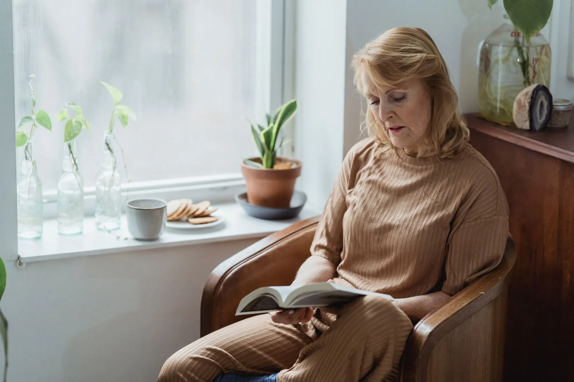 An older woman reading in an armchair beside a sunny window, with tea and biscuits nearby