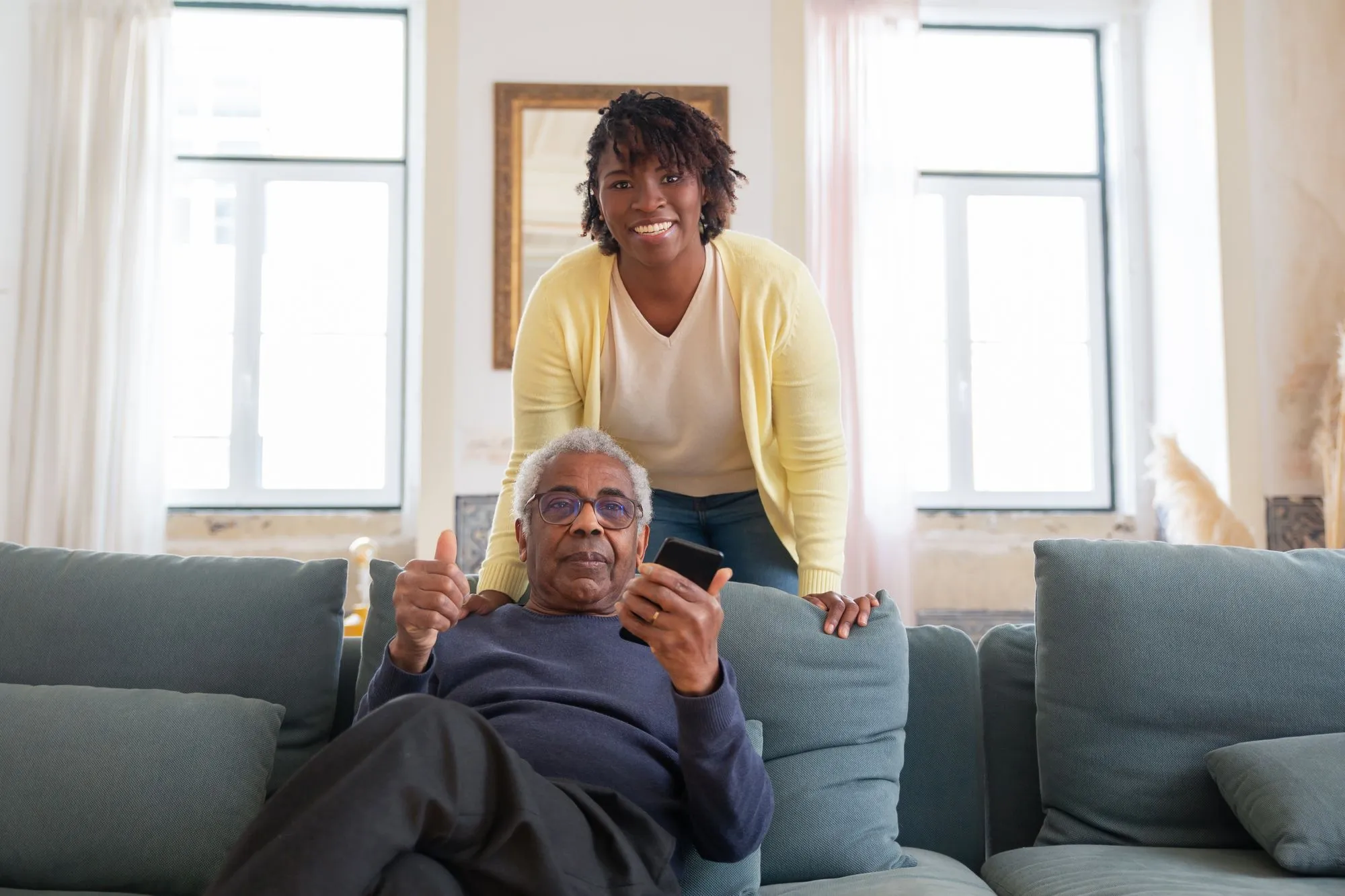 An older Black man relaxing on a sofa with a smiling Black caregiver nearby
